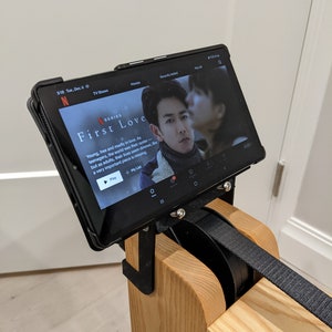 May include: A black tablet with a screen displaying the Netflix series "First Love" is mounted on a wooden stand. The tablet is attached to the stand with a black strap.