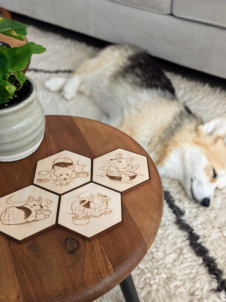 Op de afbeelding: Vier zeshoekige houten onderzetters met een schattig cartoon-hondendesign. Elke onderzetter toont een andere illustratie van de hond die geniet van een kom ramen. De onderzetters zijn in een cirkel op een houten tafel gerangschikt.