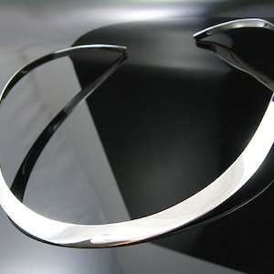 May include: A silver, curved, minimalist necklace. The necklace is made of a single, smooth piece of metal.