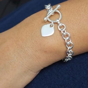 May include: A silver-toned chain link bracelet featuring a toggle clasp closure and a smooth, heart-shaped pendant charm. The bracelet is constructed from a series of interconnected circular links, creating a continuous loop around the wrist.
