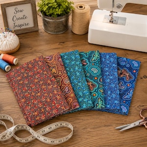 May include: Six folded fabric swatches featuring intricate patterns of flowers, dragonflies, and dots in shades of red, orange, teal, and blue. The collection is arranged in a fan shape alongside a white measuring tape, a sewing machine, a spool of thread, and a framed sign with the text Sew Create Inspire.