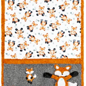 May include: A baby blanket with a white background covered in cartoon foxes in various poses. The blanket has an orange border and a gray panel with a fox applique. A plush fox toy is at the bottom.