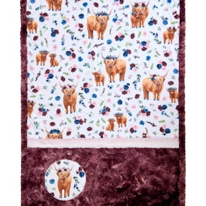 May include: A plush, rectangular blanket with a white background featuring brown Highland cows wearing floral crowns, interspersed with blue and pink flowers. The blanket has a dark burgundy border and a matching plush bottom section with a circular cow graphic.