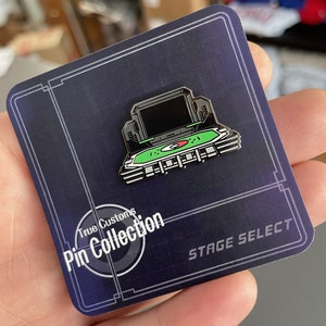 May include: A black and green enamel pin featuring a retro video game console with the text "Stage Select" and "True Customs Pin Collection".