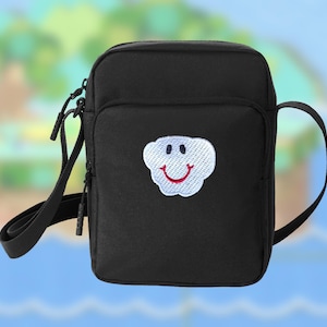 Puede incluir: Bolso bandolera negro con un parche de nube blanca con una cara sonriente. El bolso tiene un compartimento principal con cremallera, un bolsillo frontal y una correa ajustable para el hombro. El fondo es una imagen borrosa de una isla tropical.