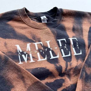 May include: A black and brown tie-dye sweatshirt with the word "MELEE" printed in white on the front. The word "MELEE" is written in a stylized font with Japanese characters above it.
