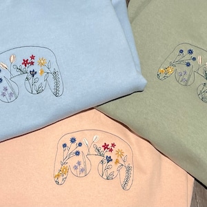 Puede incluir: Tres sudaderas con capucha de diferentes colores, azul claro, verde claro y melocotón, con un diseño bordado de un mando de videojuego con flores en su interior.