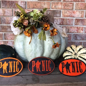 May include: Three oval-shaped black and orange embroidered patches with the word "PANIC" in white lettering and a silhouette of a person holding a gun. The patches are on a wooden surface in front of a brick wall.