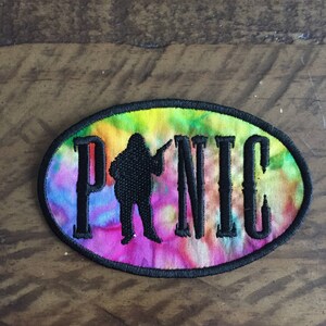 May include: A tie-dye fabric patch with a black silhouette of a person holding a rifle and the word "Panic" in black embroidery.