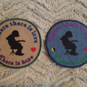 May include: Two embroidered patches with a silhouette of a person sitting on a chair holding a guitar. The patches are beige and blue with green and blue stitching. The text on the patches reads "Where there is love there is hope".