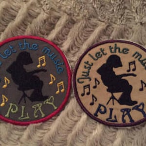May include: Two round embroidered patches with the text "Just let the music play" and a silhouette of a person playing music. One patch is on a gray background with red, yellow, and blue embroidery. The other patch is on a tan background with purple, yellow, and blue embroidery.