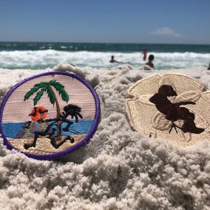 May include: Two embroidered patches on a sandy beach. One patch features a palm tree and a person with a drink in their hand. The other patch is a brown silhouette of a person on a white background.