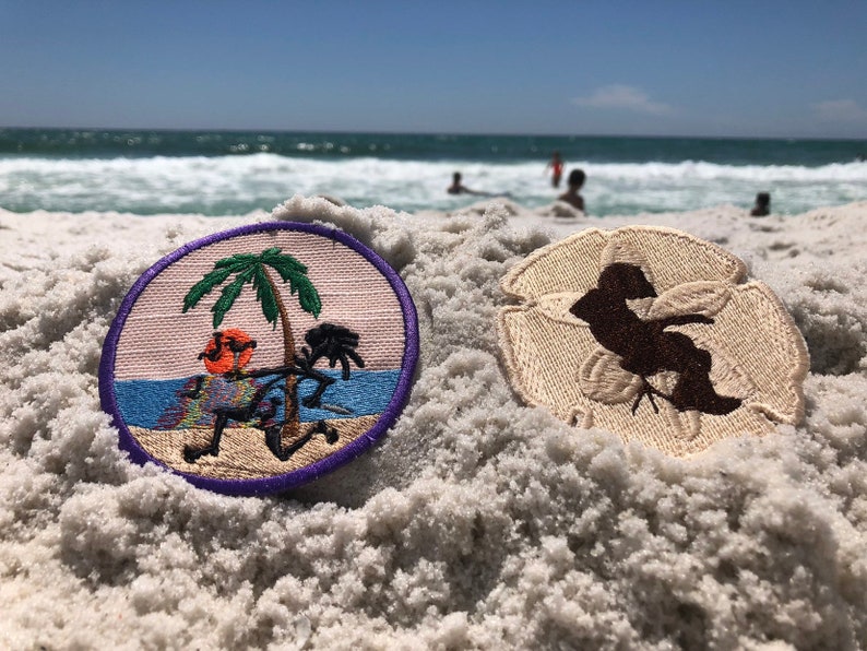 May include: Two embroidered patches on a sandy beach. One patch features a palm tree and a person with a drink in their hand. The other patch is a brown silhouette of a person on a white background.