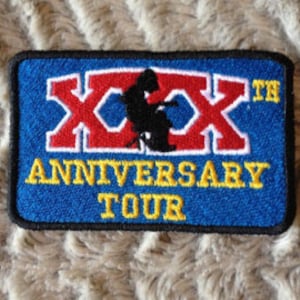 May include: A blue and yellow embroidered patch with the text "XXXth Anniversary Tour" and a silhouette of a person sitting in a chair.