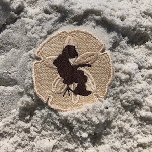 May include: A beige sand dollar with a brown embroidered silhouette of a person sitting in a chair. The sand dollar is on a white sandy beach.