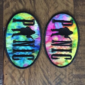 May include: Two tie-dye fabric patches with black embroidered outlines. The patches are oval shaped and feature the word "Panic" in black lettering with a silhouette of a person in the middle of the word.