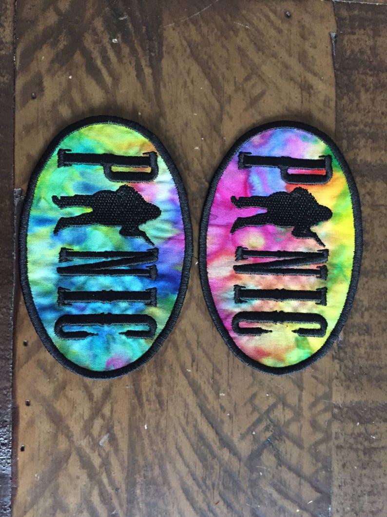May include: Two tie-dye fabric patches with black embroidered outlines. The patches are oval shaped and feature the word "Panic" in black lettering with a silhouette of a person in the middle of the word.