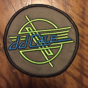 May include: A gray circular patch with a black border. The patch features a green and blue design with the text "J.J. Cale" in blue.