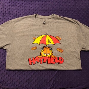 May include: Gray t-shirt with a cartoon cat wearing a yellow and red umbrella with the letters "LA" on it. The cat is surrounded by small orange and brown rectangles. The text "HATFIELD" is printed below the cat.