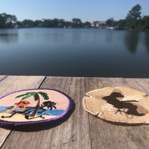 May include: Two embroidered patches on a wooden surface. One patch features a palm tree and a cartoon bird on a beach. The other patch features a silhouette of a bird on a sand dollar.