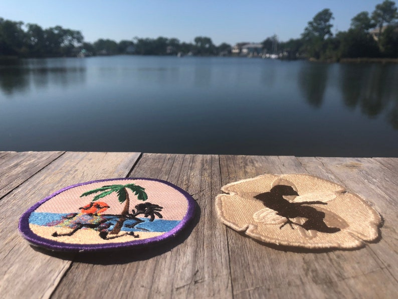 May include: Two embroidered patches on a wooden surface. One patch features a palm tree and a cartoon bird on a beach. The other patch features a silhouette of a bird on a sand dollar.