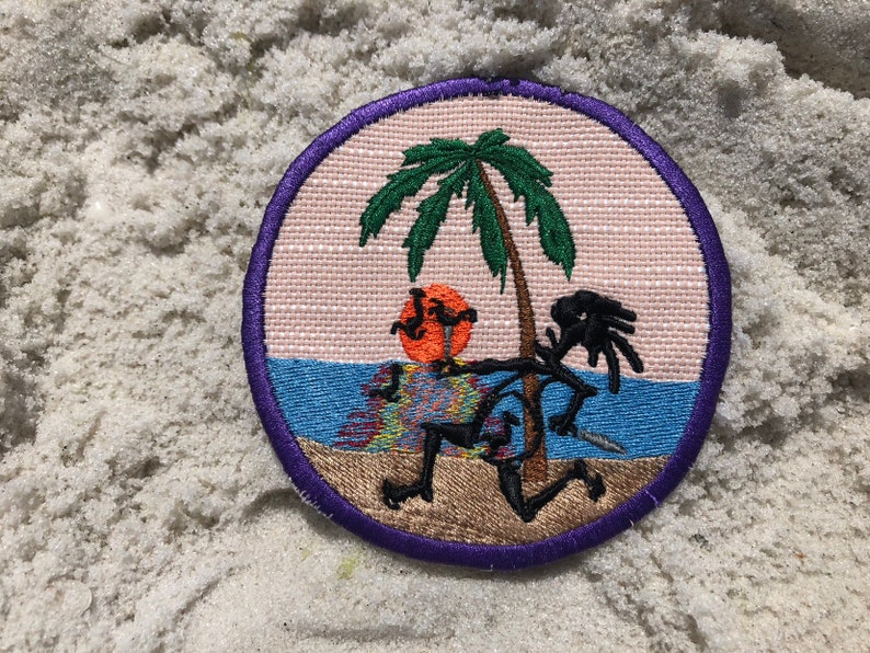May include: A round embroidered patch with a purple border. The patch features a cartoon stick figure with a palm tree and a setting sun in the background. The stick figure is wearing a colorful patterned shirt and shorts.