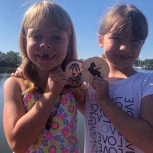 May include: Two young girls are smiling and holding sand dollar ornaments. The girl on the left is wearing a floral swimsuit and a necklace. The girl on the right is wearing a white t-shirt with the word "LOVE" repeated in black.