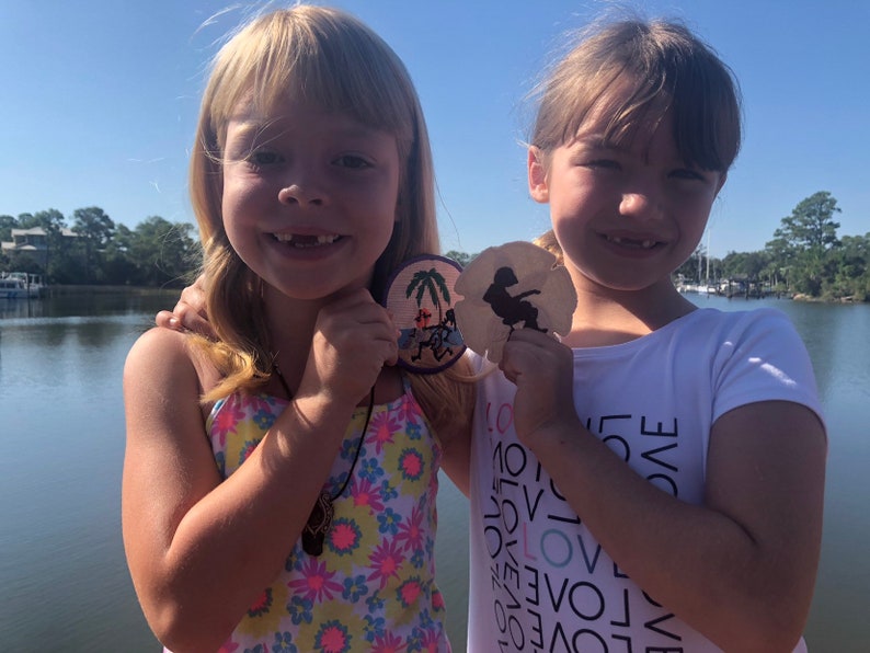 May include: Two young girls are smiling and holding sand dollar ornaments. The girl on the left is wearing a floral swimsuit and a necklace. The girl on the right is wearing a white t-shirt with the word "LOVE" repeated in black.