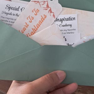 May include: A sage green envelope containing several paper cards and a tag. The tag features a mountain graphic and the text Heart Is In Highlands. Other cards display text including Special Hayride in the and Inspiration Cranberry.