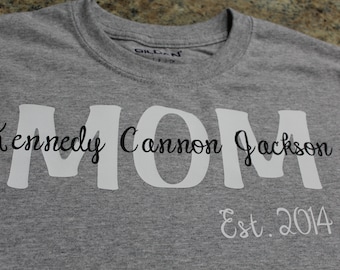 Può includere: Una maglietta grigia girocollo con la parola "MOM" a grandi lettere bianche. Sotto la parola "MOM" c'è il nome "Kennedy Cannon Jackson" in corsivo nero. Il testo "Est. 2014" è sotto il nome.