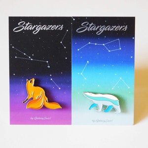 May include: Two enamel pins on a card with a starry night sky background. The pins are part of a set called "Stargazers" by Galaxy Snail. The left pin features a yellow fox looking up at the stars. The right pin features a white polar bear with blue accents walking across the sky.