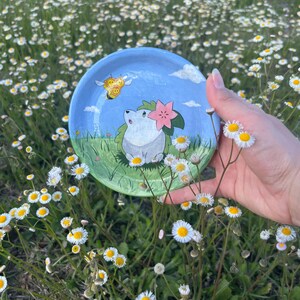 Pode incluir: Um pequeno prato de cerâmica pintado à mão com um ouriço de desenho animado com uma flor rosa e uma abelha amarela contra um fundo de céu azul. O prato é segurado em frente a um campo de flores brancas.