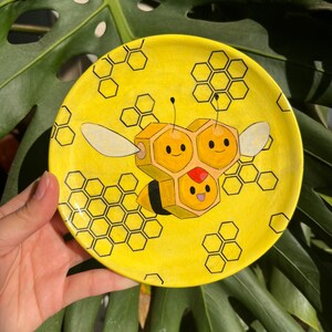 May include: A yellow ceramic plate featuring a cartoon bee design. The bee is composed of honeycomb shapes with a smiling face and wings. Black honeycomb patterns surround the bee. The plate is held against a backdrop of green leaves.