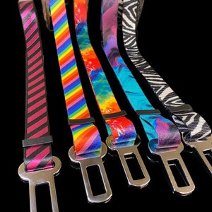 May include: Five colorful dog seatbelt harnesses with silver metal clips. The harnesses are striped, tie-dye, and zebra patterned.