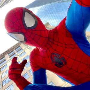 Könnte beinhalten: Eine Person in einem Spider-Man-Kostüm, rot und blau mit einem Spinnennetz-Muster. Die Maske hat weiße Augenlinsen und ein schwarzes Spinnenemblem auf der Brust. Im Hintergrund sind hohe Gebäude und ein blauer Himmel zu sehen.