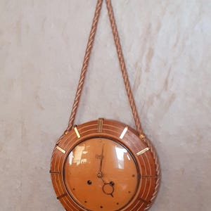 Puede incluir: Un reloj de pared redondo de madera con una esfera marrón claro y un marco de madera marrón oscuro. El reloj tiene detalles dorados y está suspendido por una cuerda. El reloj está colgado en una pared de color claro.