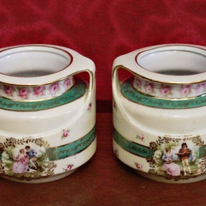 May include: Two white porcelain vases with green and gold trim. Each vase features a romantic scene with two figures and a floral design. The vases have two handles on each side.