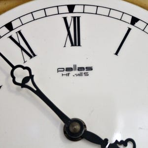 May include: A white clock face with black Roman numeral hour markers and black hands. The clock face has the text "pallas HF_GES" in black.