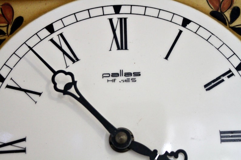 May include: A white clock face with black Roman numeral hour markers and black hands. The clock face has the text "pallas HF_GES" in black.
