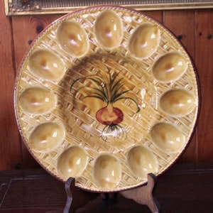 Puede incluir: Un plato redondo de cerámica para huevos con 12 hendiduras individuales. El plato está decorado con un patrón de cesta tejida y una cebolla pintada con hojas verdes.