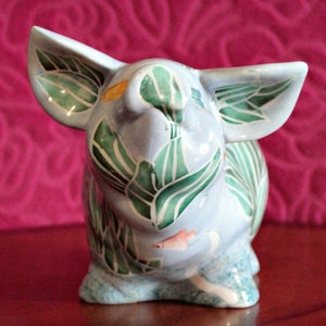 Può includere: Un salvadanaio in ceramica blu con disegni di foglie verdi e bianche e un piccolo pesce arancione dipinto sul muso. Il salvadanaio è seduto su una superficie rosa.