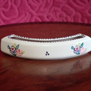 Puede incluir: Macetero de cerámica blanca con forma curva y diseños florales en rosa, morado y verde. El macetero tiene un pequeño borde alrededor del borde superior.