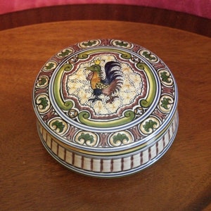 May include: A round ceramic trinket box with a colorful rooster design. The box has a white background with green, brown, and blue accents. The rooster is depicted in the center of the lid, with a yellow crown on its head.