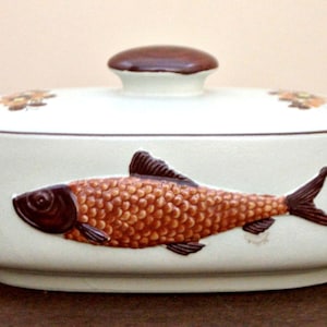 Puede incluir: Una cacerola de cerámica blanca con tapa marrón y un diseño de pez marrón. El pez tiene escamas naranjas y un cuerpo marrón.