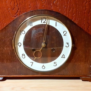 May include: A vintage wooden mantel clock with a white clock face and black numerals. The clock has a curved top and a dark brown finish. The clock hands are gold-coloured. The clock is a classic decorative piece.