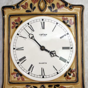 May include: A ceramic wall clock with a yellow and brown frame and floral design. The clock face is white with black Roman numeral markings and the words "Quartz" and "Paliss" printed on it.