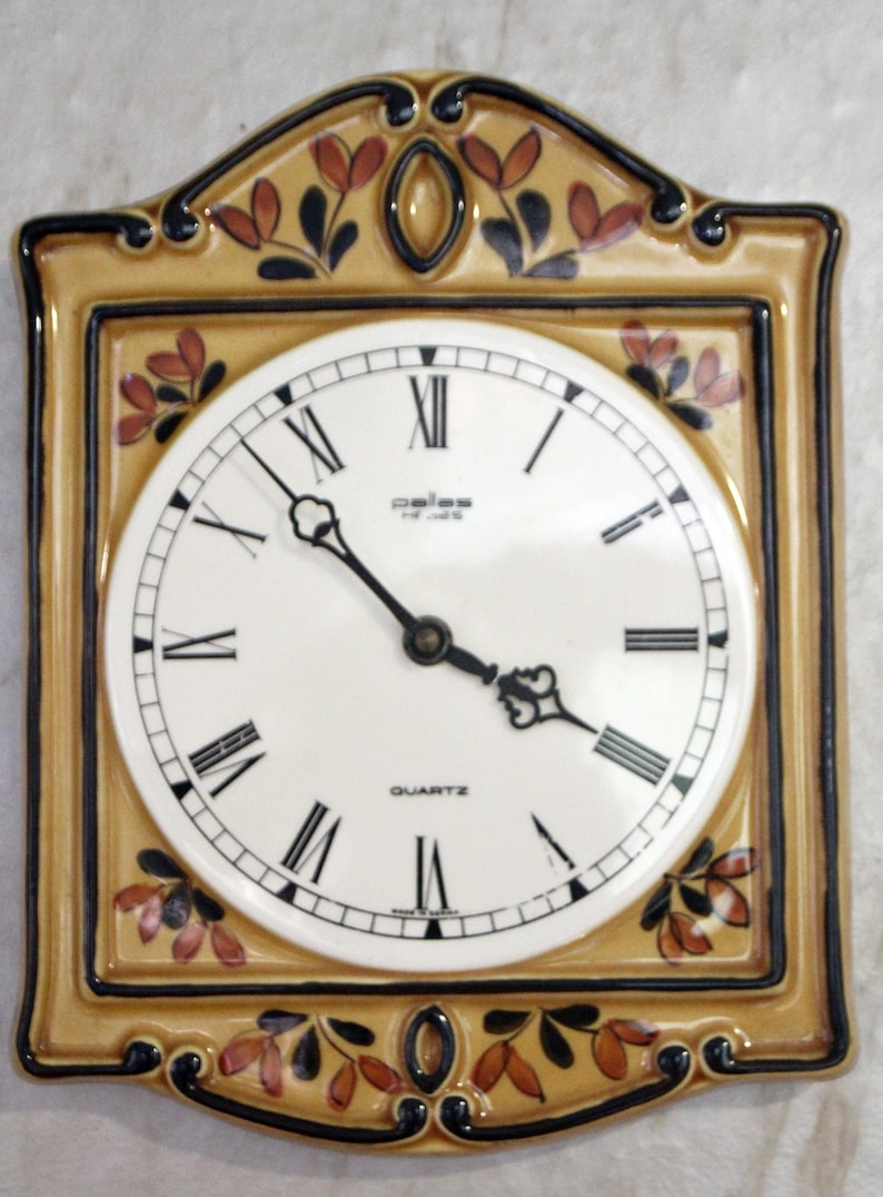 May include: A ceramic wall clock with a yellow and brown frame and floral design. The clock face is white with black Roman numeral markings and the words "Quartz" and "Paliss" printed on it.