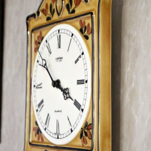 May include: A ceramic wall clock with a yellow and brown frame and a white face. The clock has black Roman numeral hour markers and a black minute hand. The clock is decorated with painted brown and orange leaves.