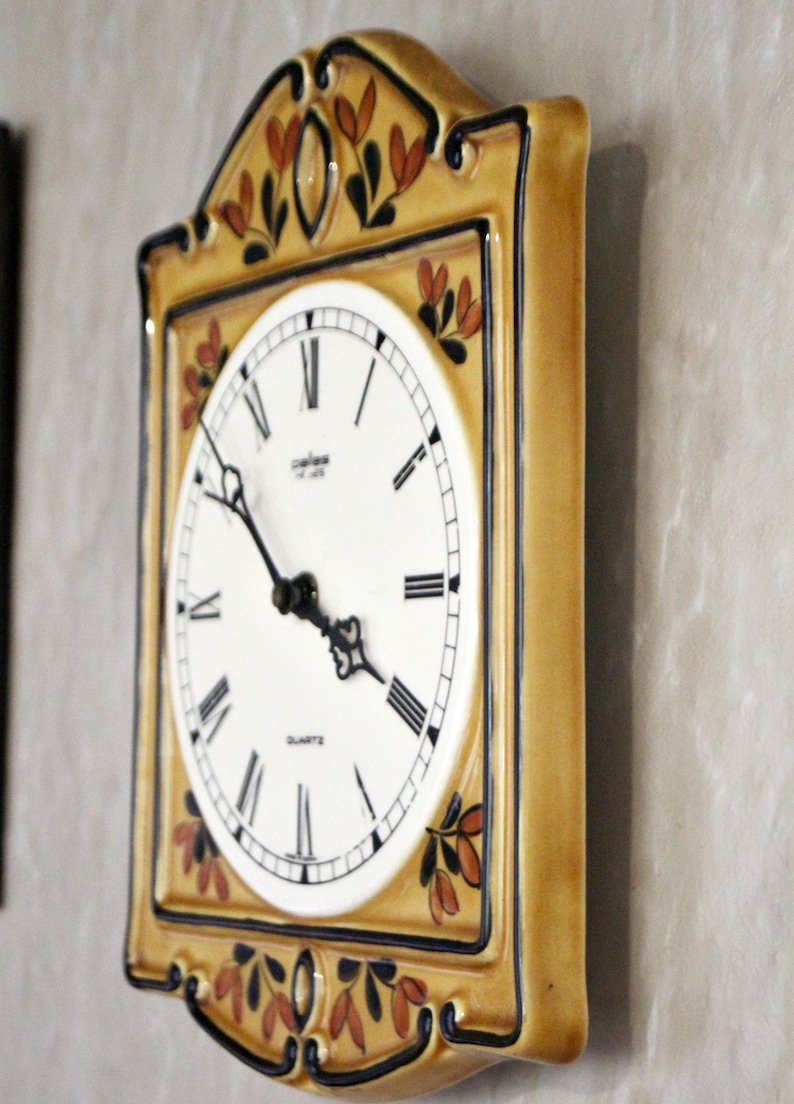 May include: A ceramic wall clock with a yellow and brown frame and a white face. The clock has black Roman numeral hour markers and a black minute hand. The clock is decorated with painted brown and orange leaves.