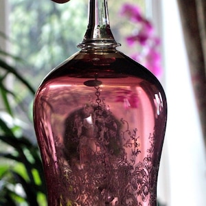 Pode incluir: Um sino de vidro transparente com uma tonalidade rosa e desenhos florais gravados intrincados. O sino tem uma borda dourada e um caule de vidro transparente.